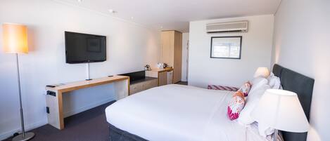A modern hotel room with a large bed, a desk with a flat-screen TV, a built-in air conditioning unit, and a window providing natural light.