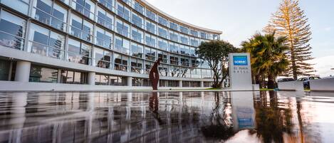 A modern hotel with a reflective pool, a sculpture, and a sign for the Scenic Hotels Te Pania Hotel.