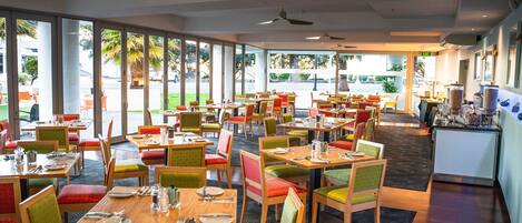 A spacious restaurant with multiple dining tables and chairs, large windows offering a view of the outdoors, and ceiling fans.
