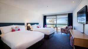 Habitación de hotel con dos camas, un escritorio, una silla, un televisor y un amplio ventanal con vista al mar.