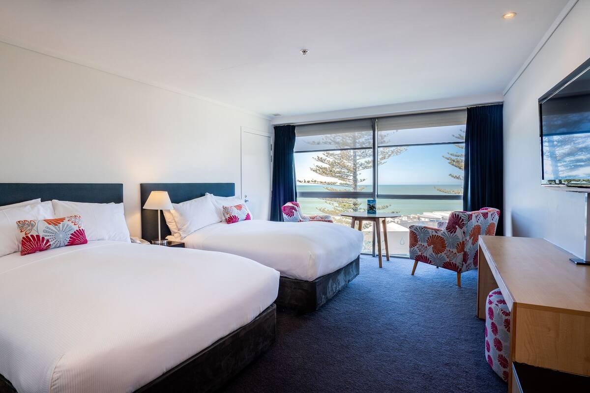 A hotel room with two beds, a desk, a chair, a TV, and a large window with a view of the sea.