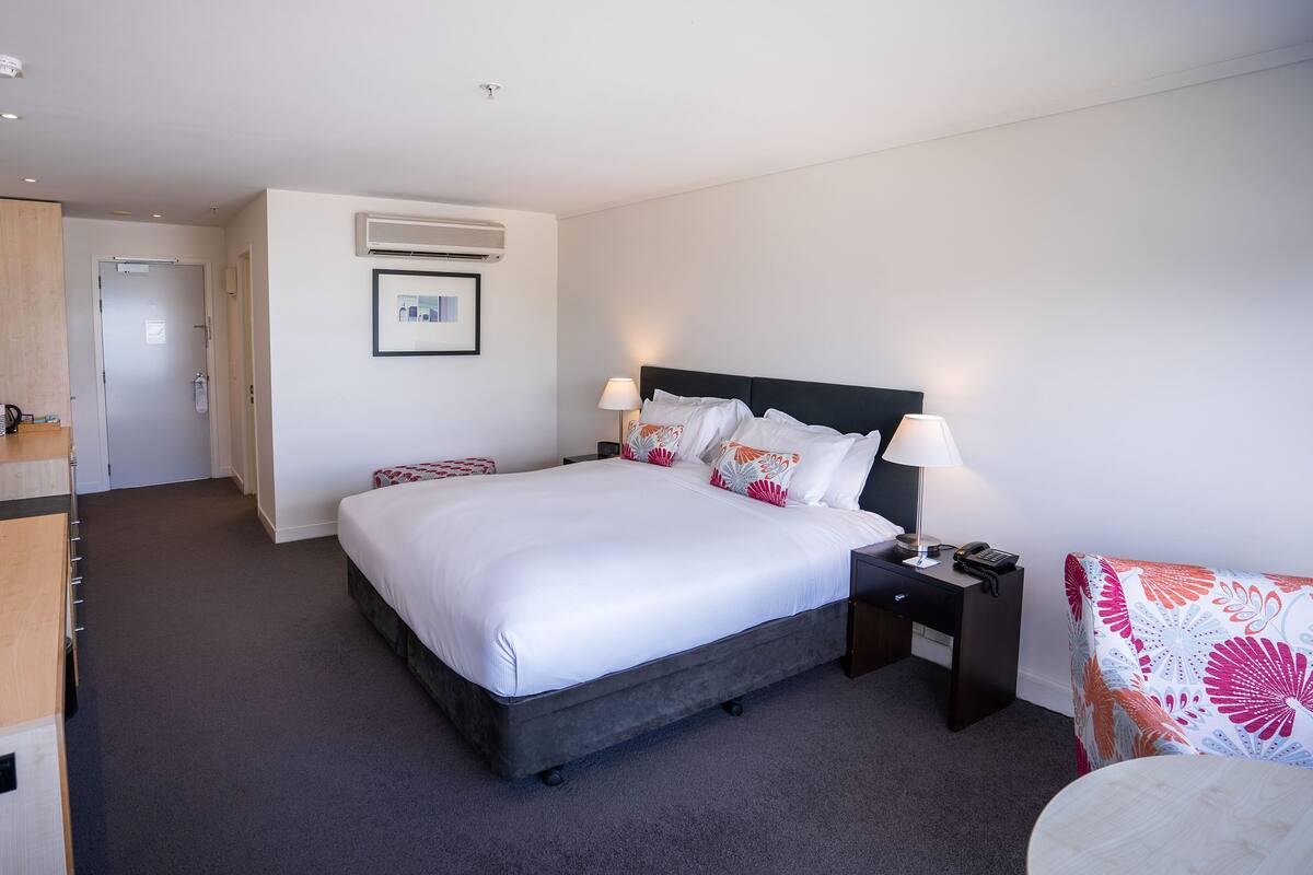 A hotel room with a large bed, bedside tables, a desk, and an air conditioning unit.