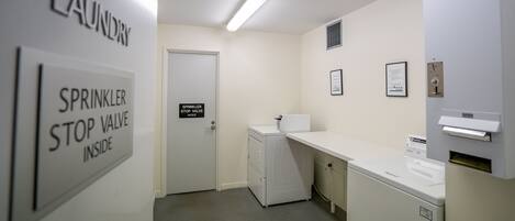 A laundry room with a white wall, a door, a white washing machine, and a dryer. There is a sign on the wall that says "Sprinkler Stop Valve Inside".