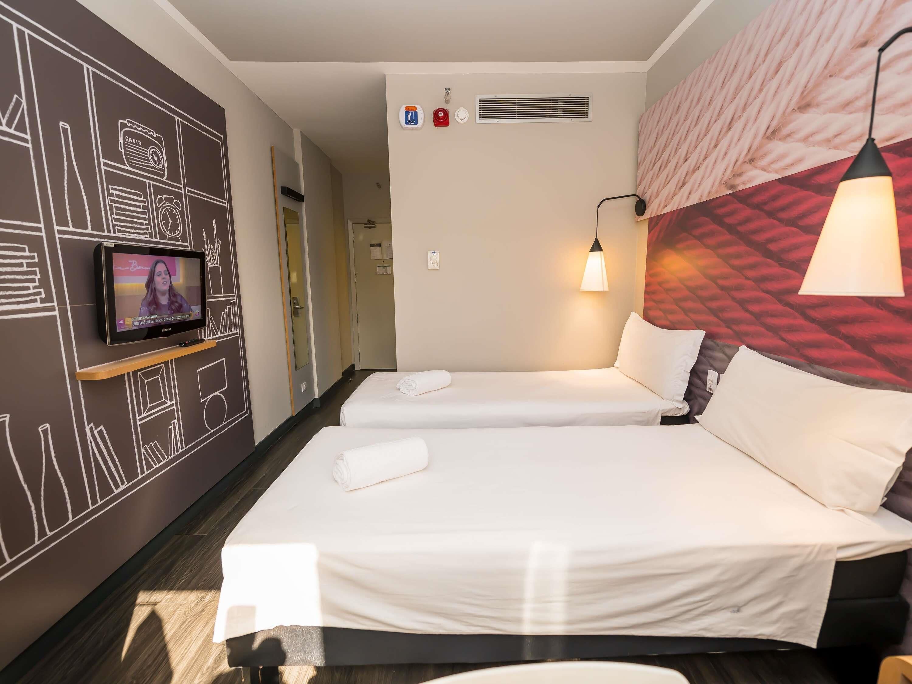 Quarto de hotel com duas camas, uma televisão na parede, cabeceira com luminária e parede com design geométrico.