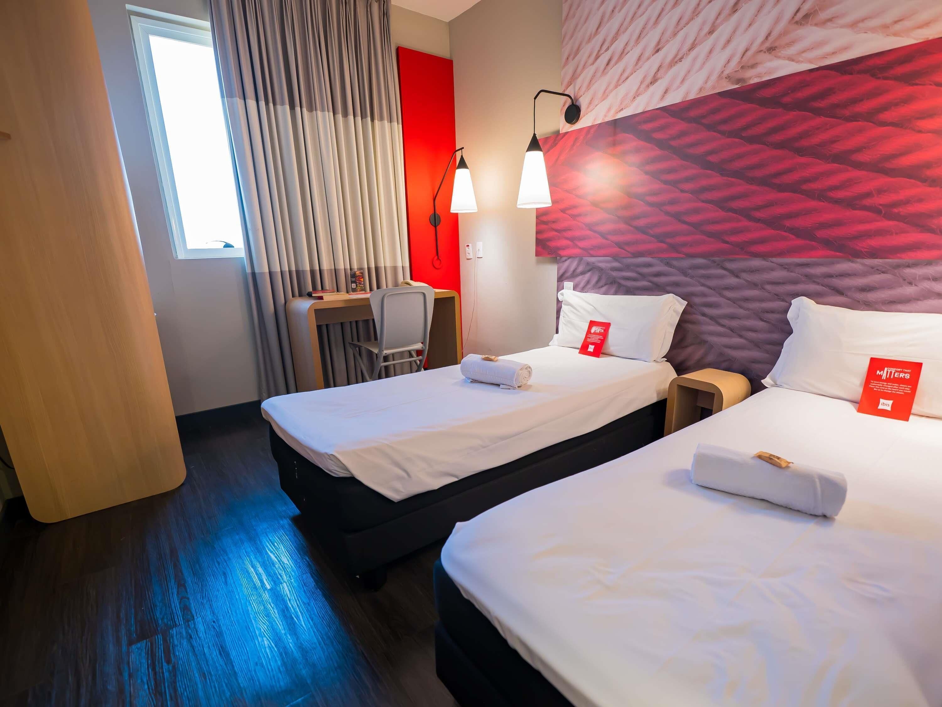 Habitación de hotel con dos camas, un escritorio, una silla, pared roja y ventana con cortinas.
