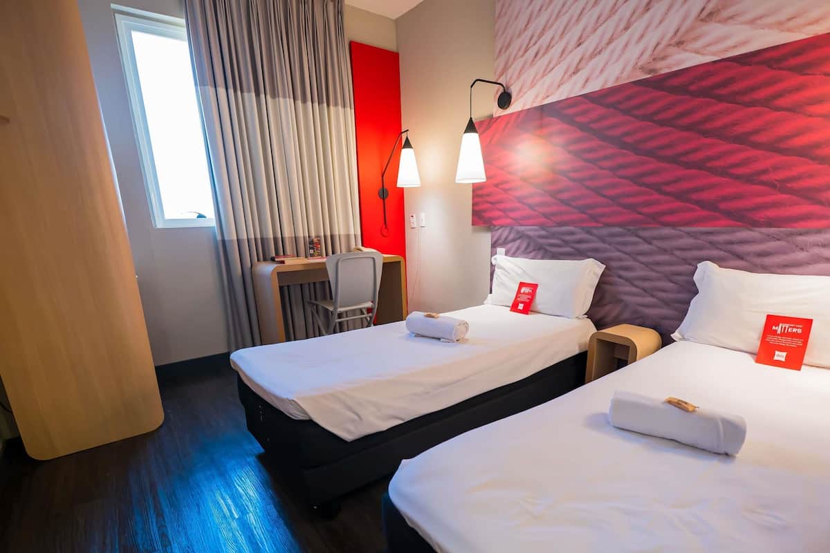 Habitación de hotel con dos camas, un escritorio y una silla. Se aprecia una ventana con cortinas, una pared de acento roja y un cabecero con textura.
