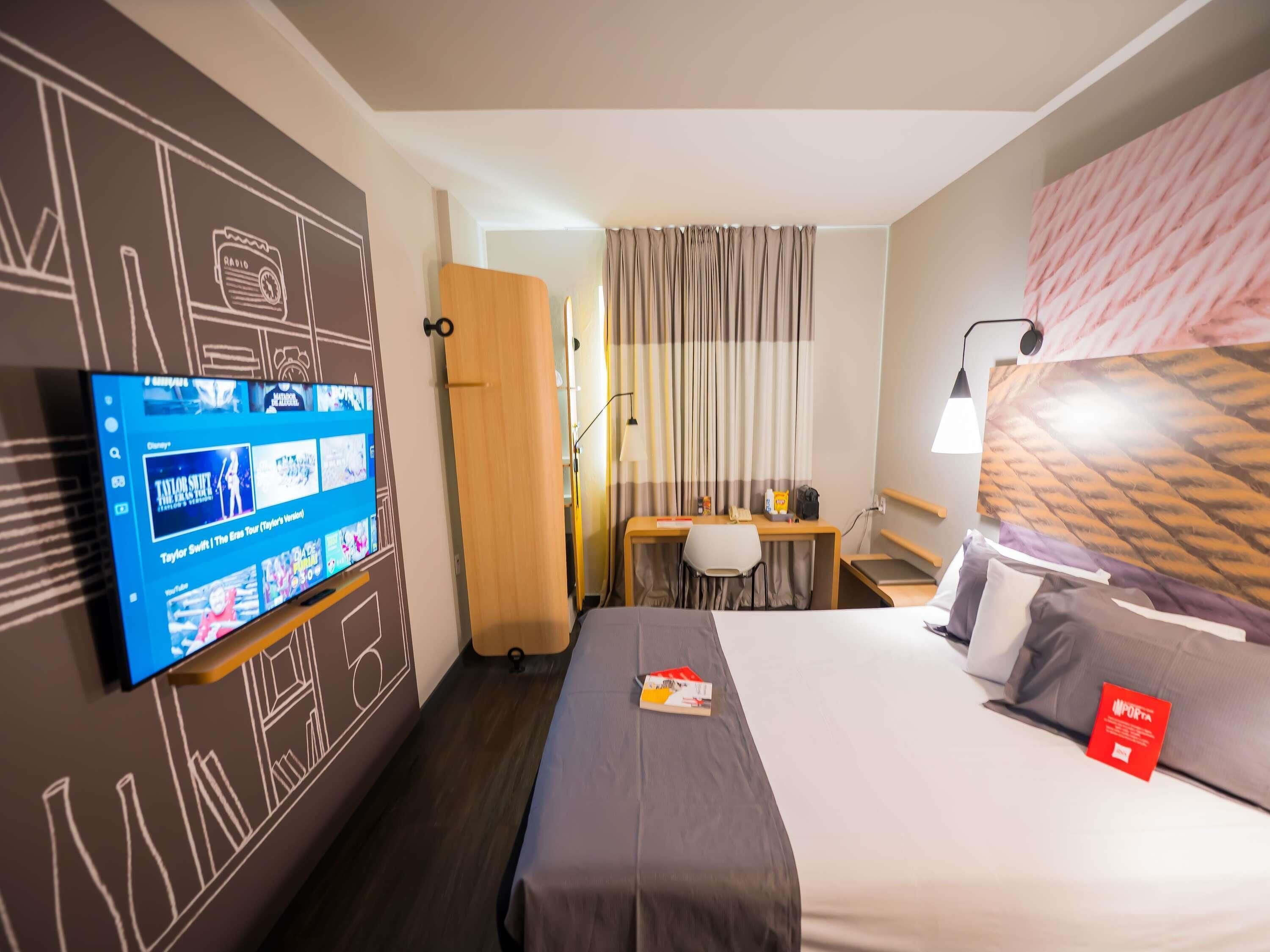 A hotel room with a bed, a desk with a chair, a TV mounted on the wall, and curtains on the window.