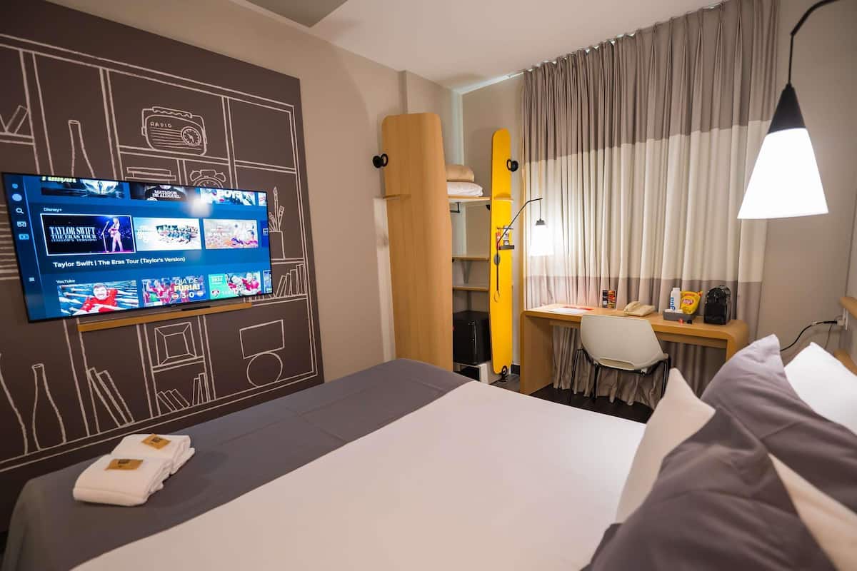 Una habitación de hotel con cama, escritorio, silla, televisión mostrando varios canales y ventana con cortinas.