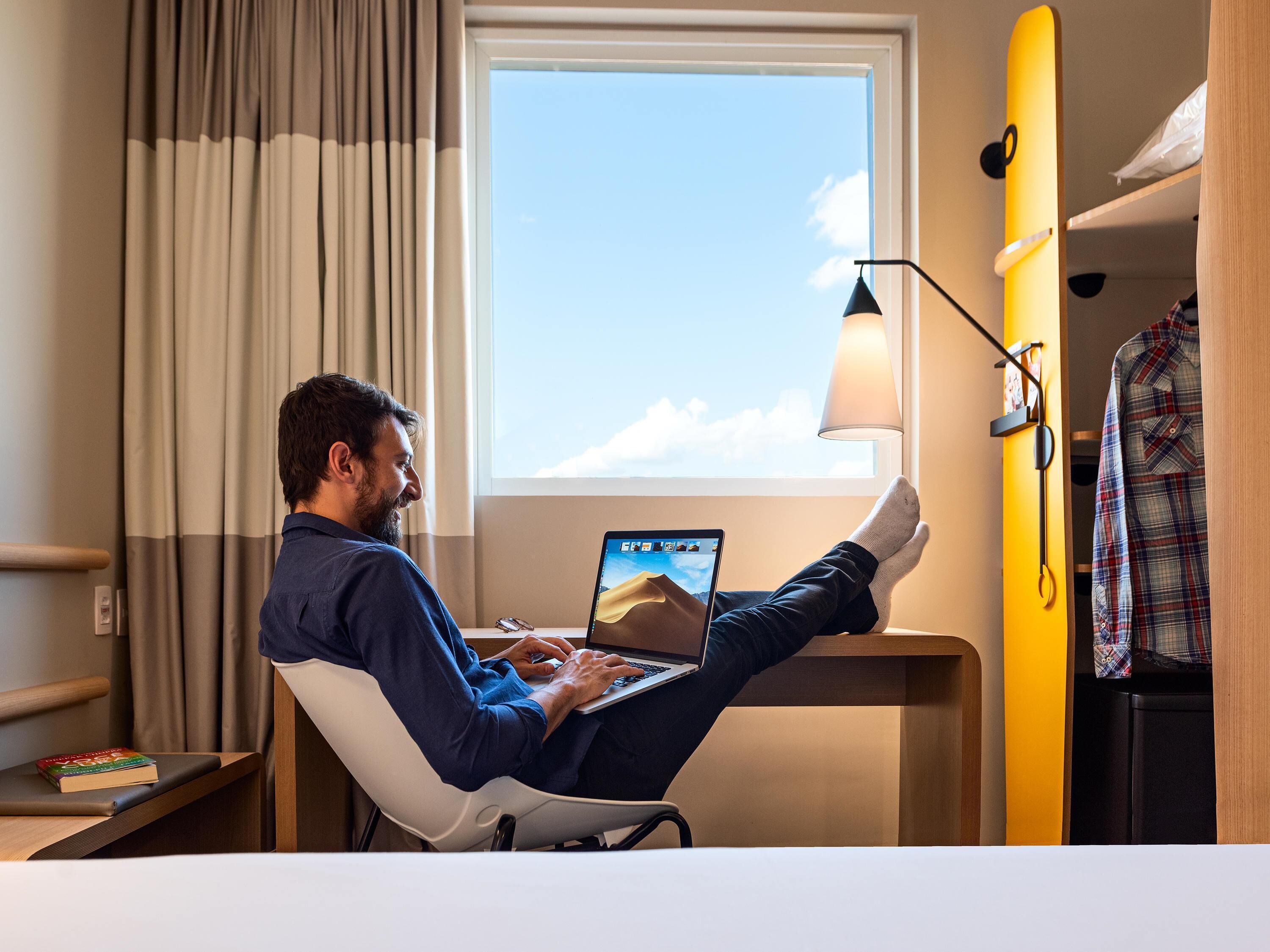 A man working on a laptop in a hotel room with a window view, a lamp, and a bed with a blanket.