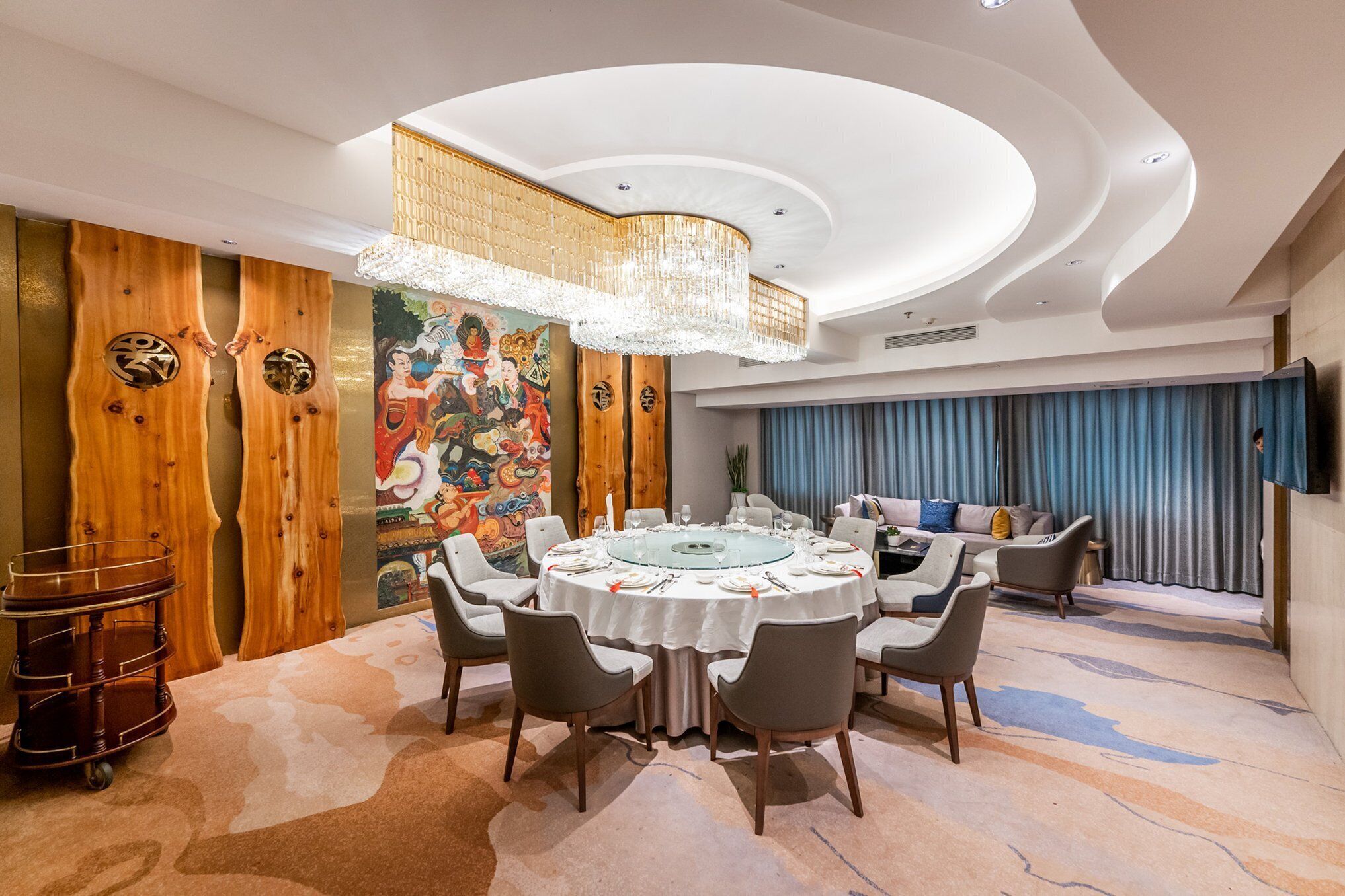 A dining area with a round table set for six, surrounded by six chairs. A chandelier hangs from the ceiling, and a large painting adorns the wall.