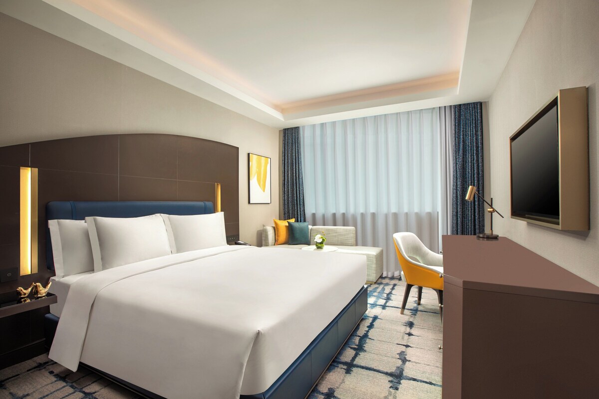 A modern hotel room with a large bed, a desk, a chair, a TV, and a window with curtains.