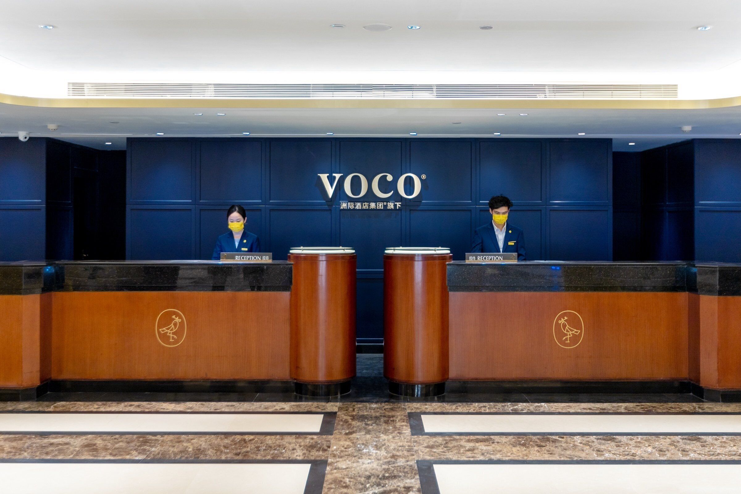 Un comptoir d’accueil avec deux réceptionnistes, un mur bleu orné du logo « VOCO » et un sol en marbre.