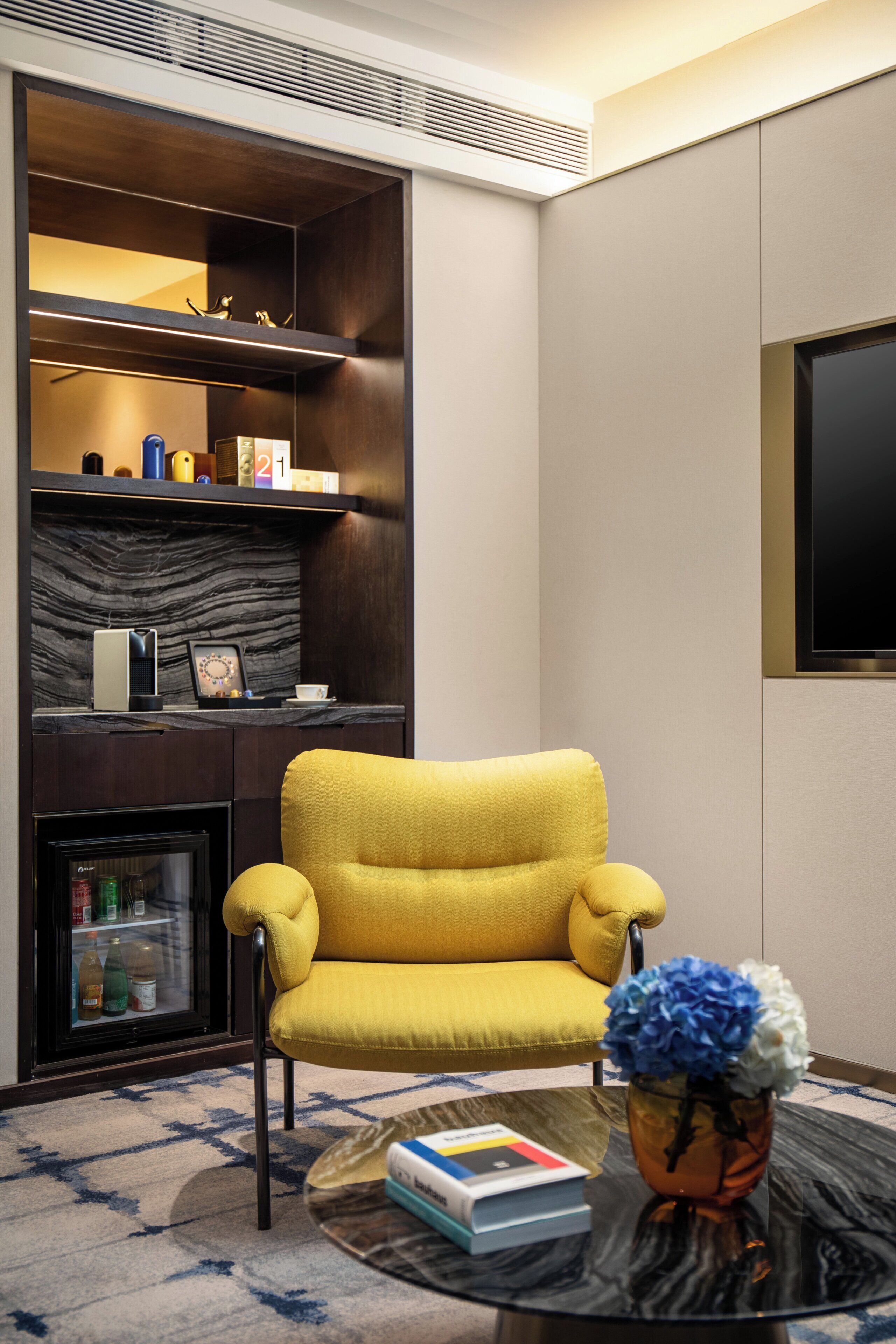 A modern living room with a yellow armchair, a glass coffee table with a book and a vase of flowers, a fireplace, and a TV mounted on the wall.