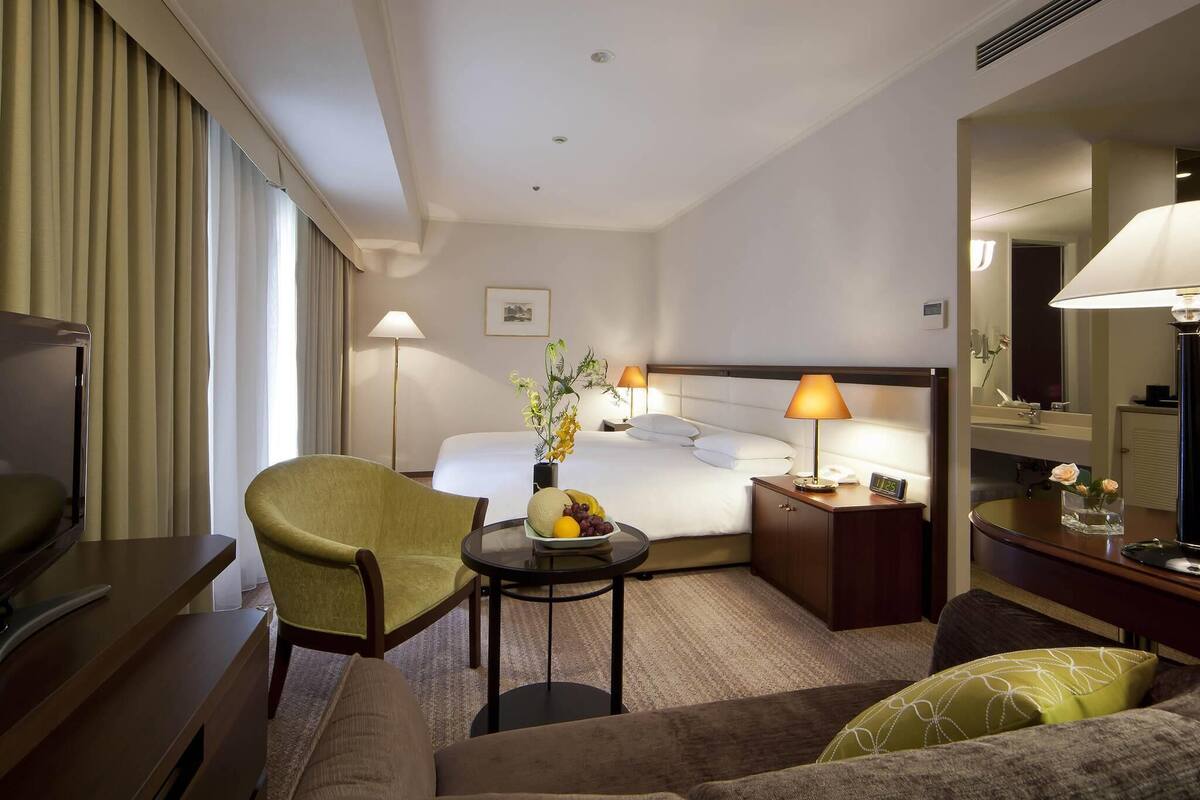 Ein Hotelzimmer mit Bett, Schreibtisch, Sessel, Sofa, Nachttisch mit Lampe, einer Uhr und einem Spiegel.