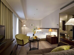 Ein Hotelzimmer mit Bett, Schreibtisch, Sessel, Sofa, Nachttisch mit Lampe, einer Uhr und einem Spiegel.