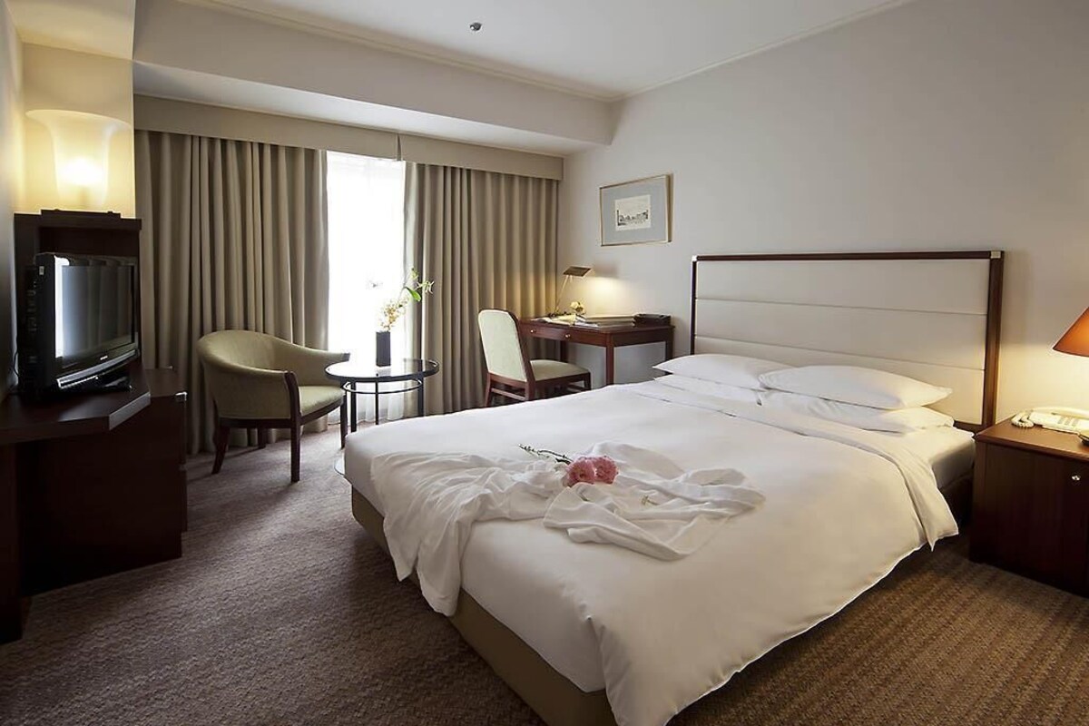 Quarto de hotel com cama, escrivaninha, cadeira, televisão e janela com cortinas.