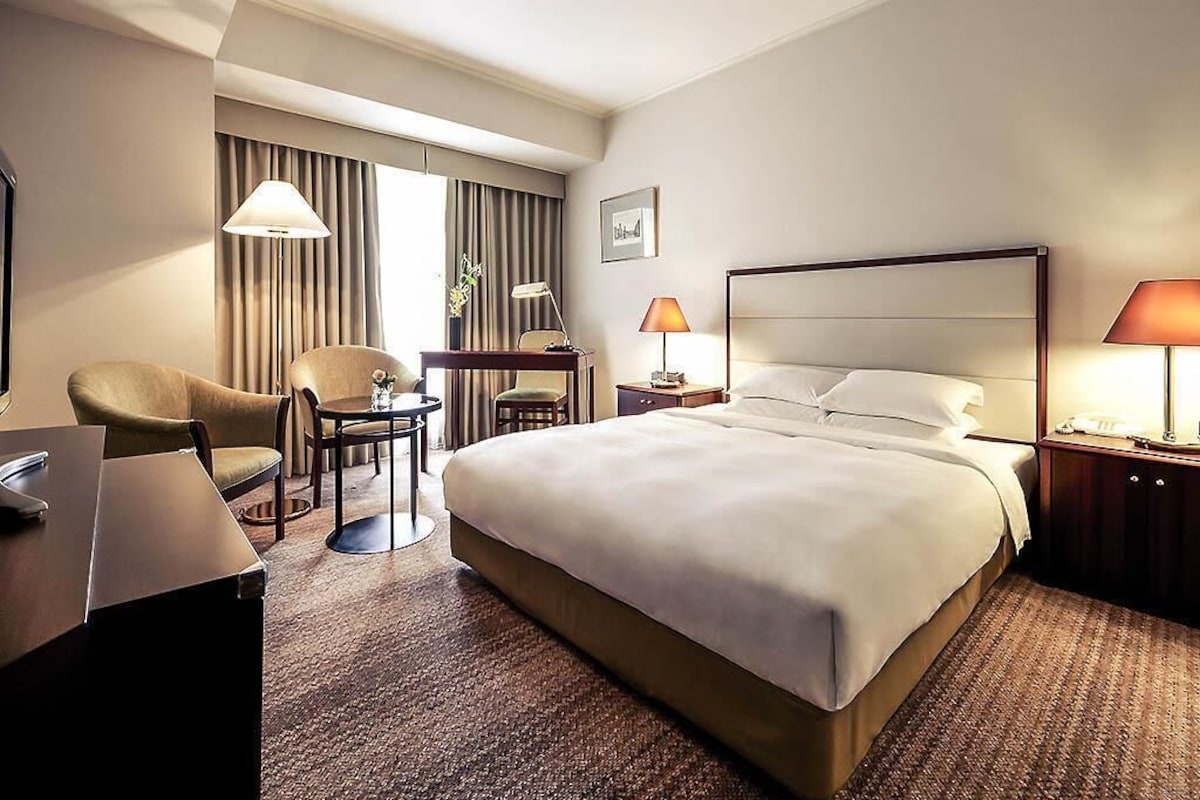Quarto de hotel com cama, duas cadeiras, escrivaninha, abajur e mesa de cabeceira.