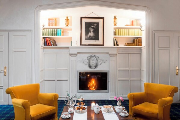 Una habitación con chimenea, dos sillones amarillos, una mesa con vajilla para té, estanterías y un retrato enmarcado.