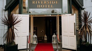 Entrada del Hotel Riverside con alfombra roja, puertas blancas y plantas en macetas.