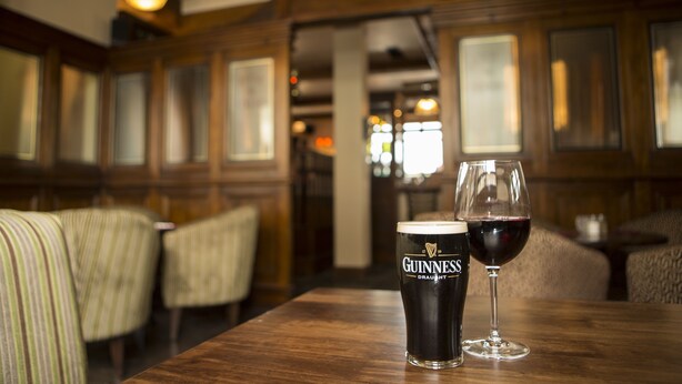 Una mesa de madera con un vaso de cerveza Guinness y un vaso de vino. El ambiente parece ser un restaurante o bar acogedor y tenuemente iluminado, con paneles de madera y asientos acolchados.