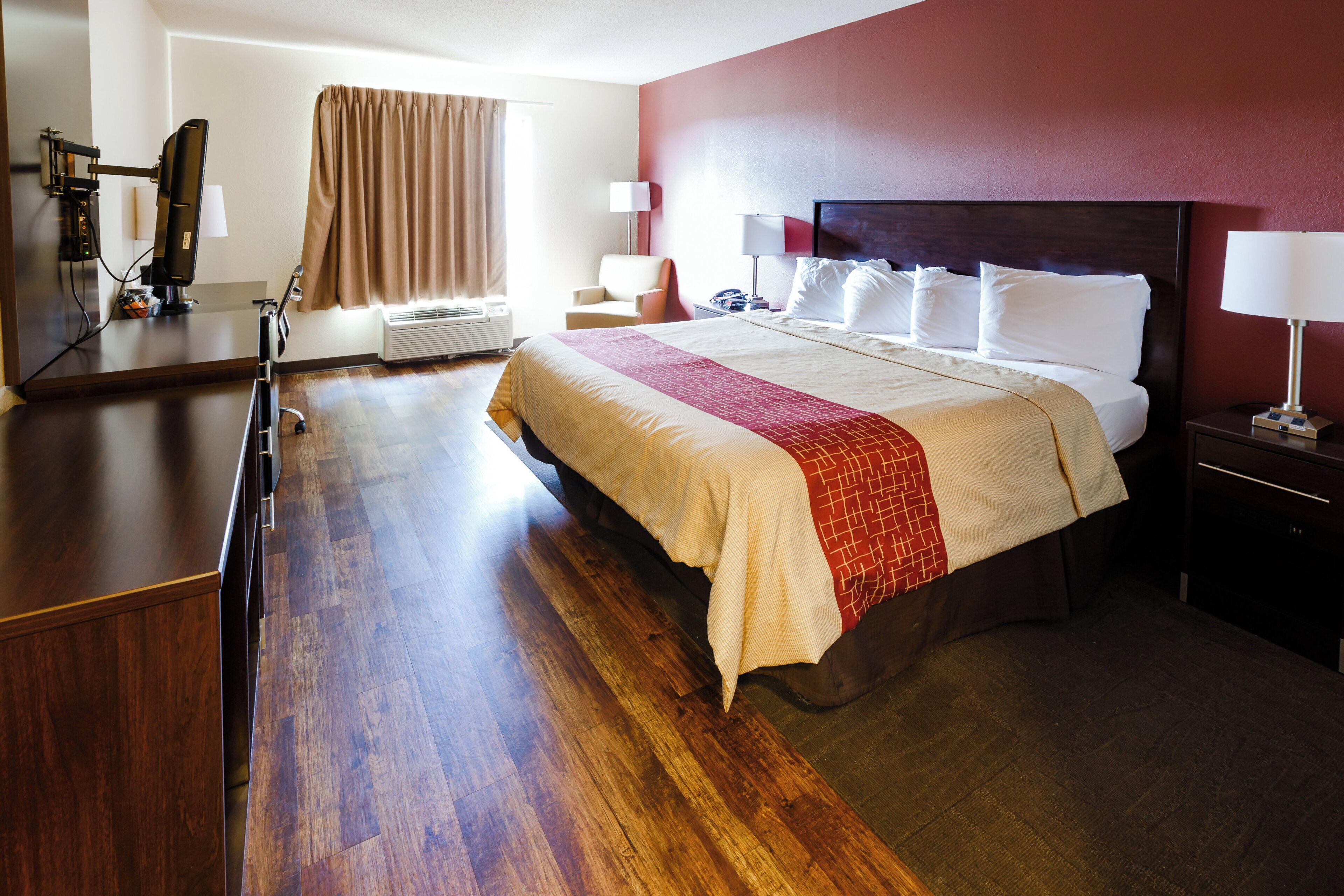 Habitación de hotel con una cama grande, un escritorio, una silla, una mesita de noche con lámpara y una ventana con cortinas.
