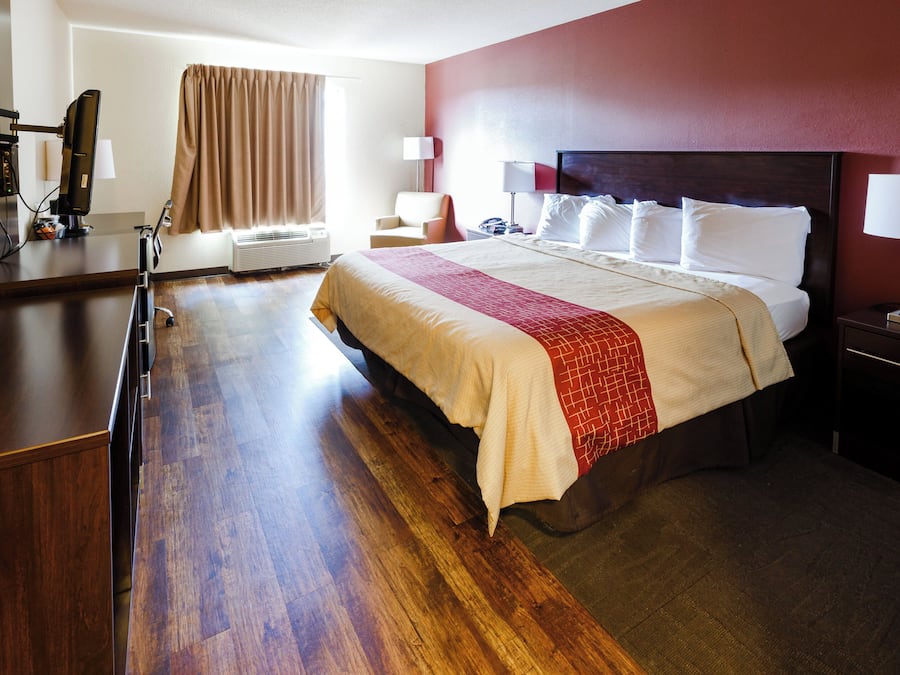 A hotel room with a large bed, a desk, a chair, a bedside table with a lamp, and a window with curtains.