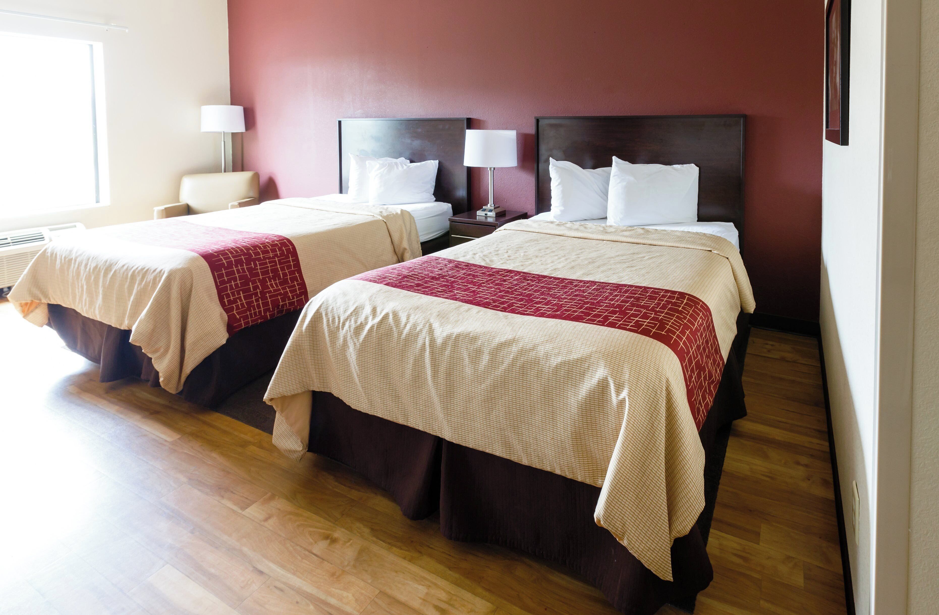 A hotel room with two beds, each with a red and beige bedspread, a nightstand with a lamp, and a window with blinds.