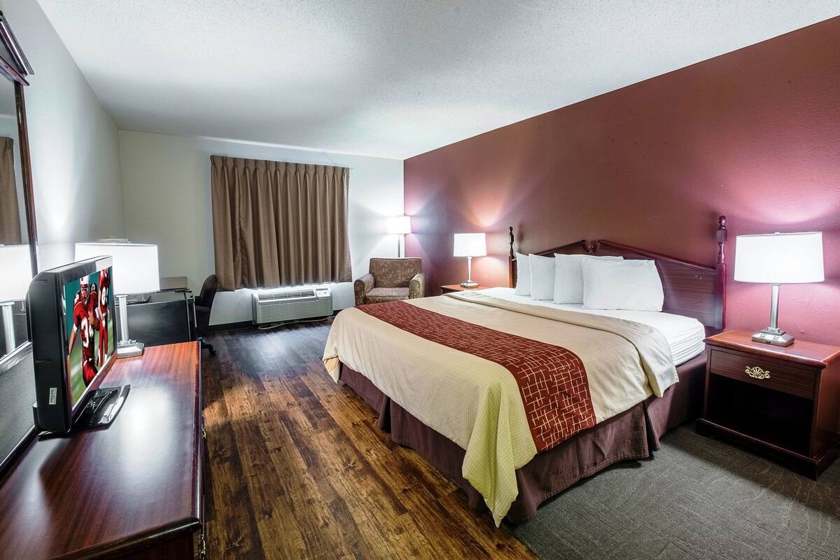 Ein Hotelzimmer mit einem Bett, einem Schreibtisch mit Fernseher, einem Sessel, einem Nachttisch mit Lampe und einem Fenster mit Vorhängen.
