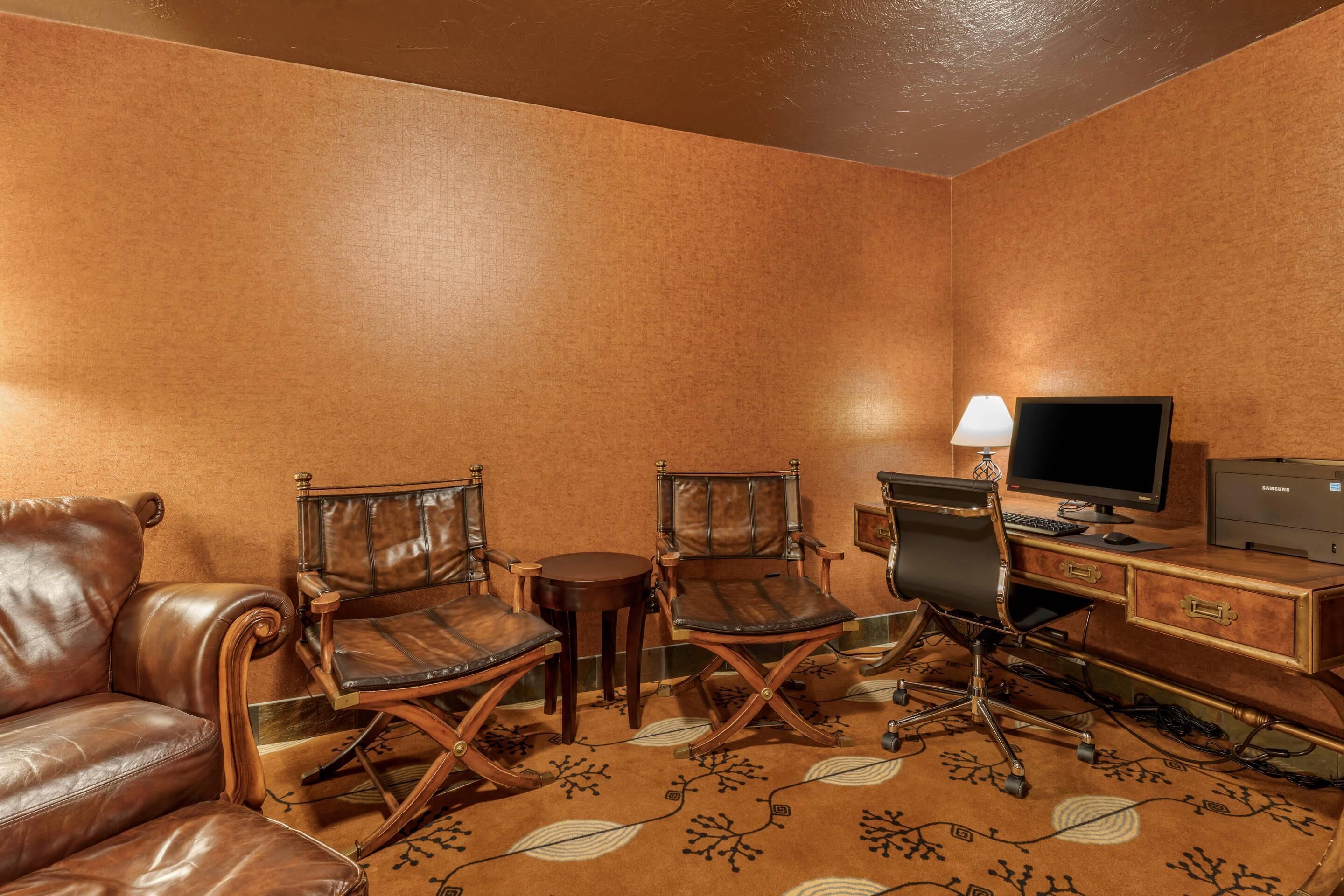 A room with a brown leather sofa, two leather chairs, a small round table, a desk with a computer and a lamp, and a patterned carpet.