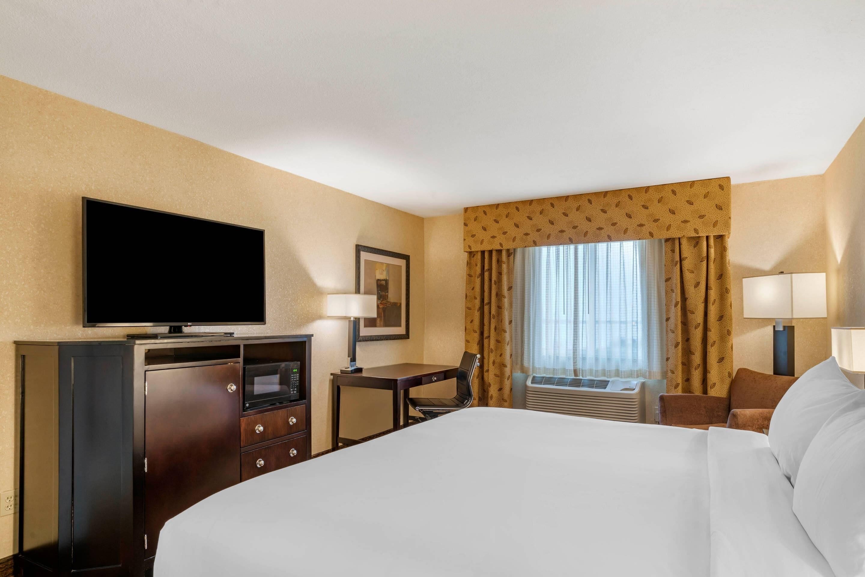 Habitación de hotel con una cama grande, un televisor de pantalla plana, un escritorio con una silla, una lámpara de escritorio y un ventanal con cortinas.