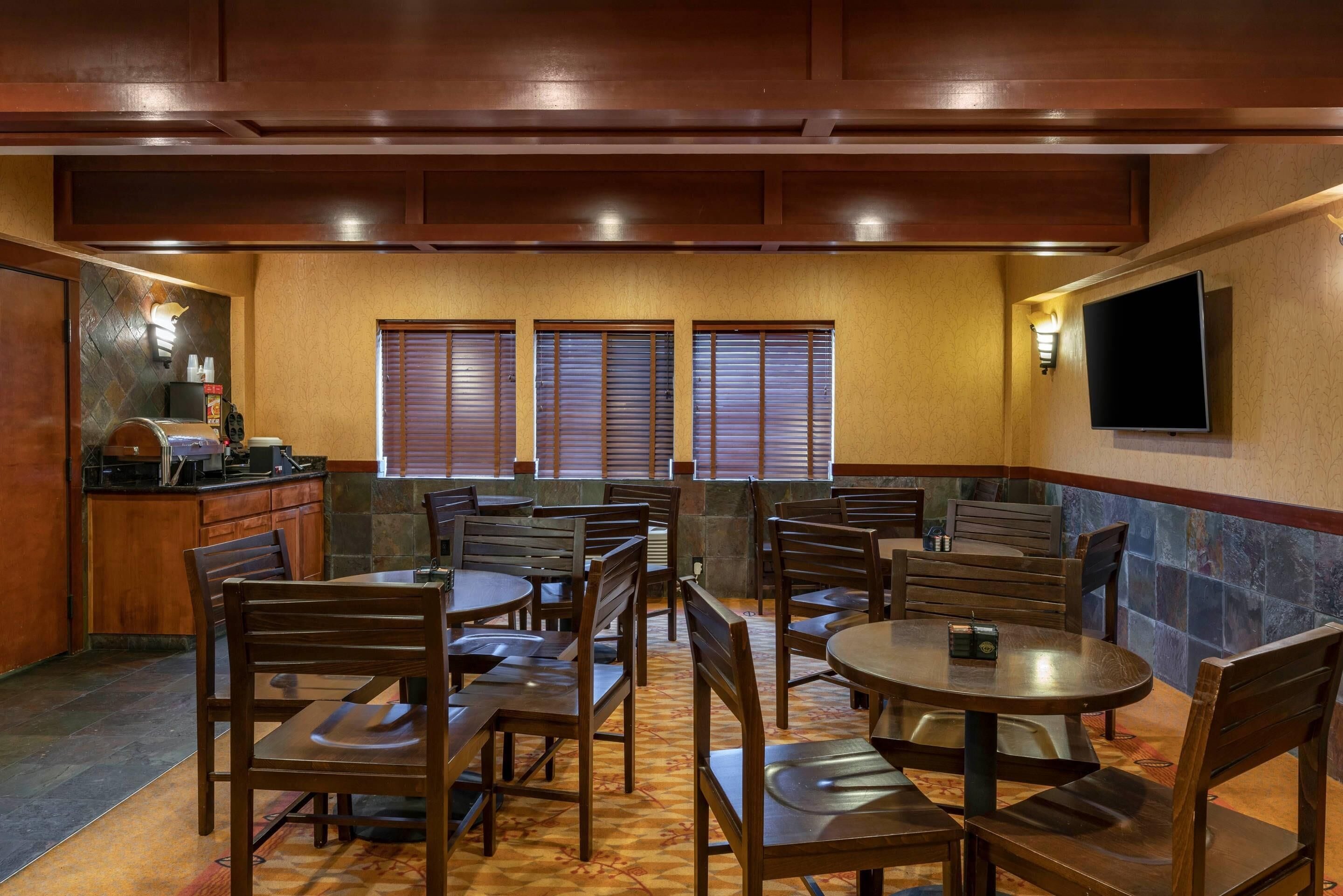A restaurant with wooden tables and chairs, a kitchen area, and a TV mounted on the wall.