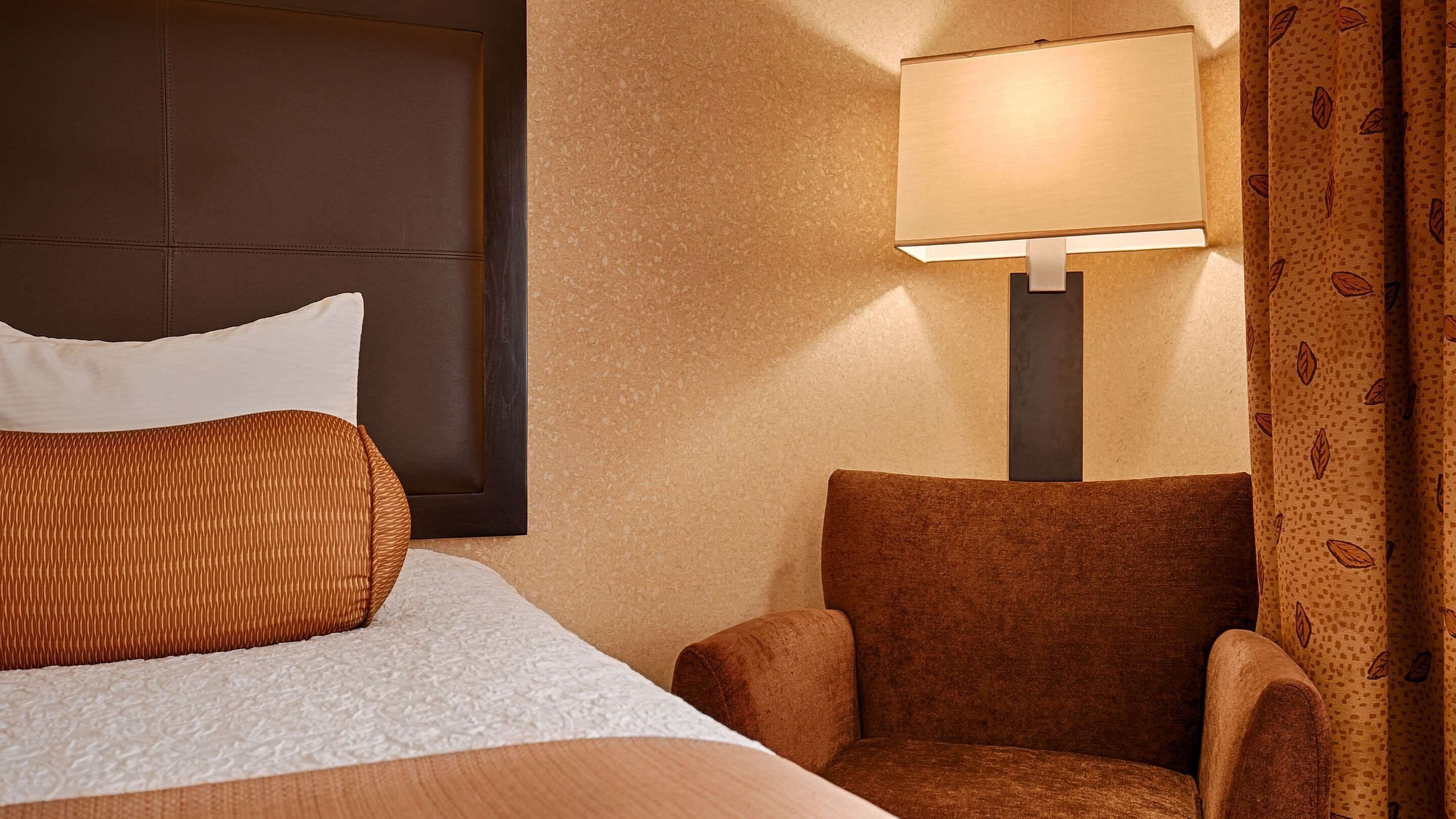 A hotel room with a bed, a brown upholstered armchair, a wall-mounted lamp, and a patterned curtain.