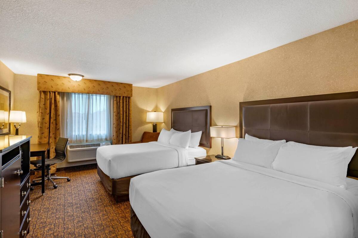 A hotel room with two beds, a desk, a chair, and a large window with curtains.