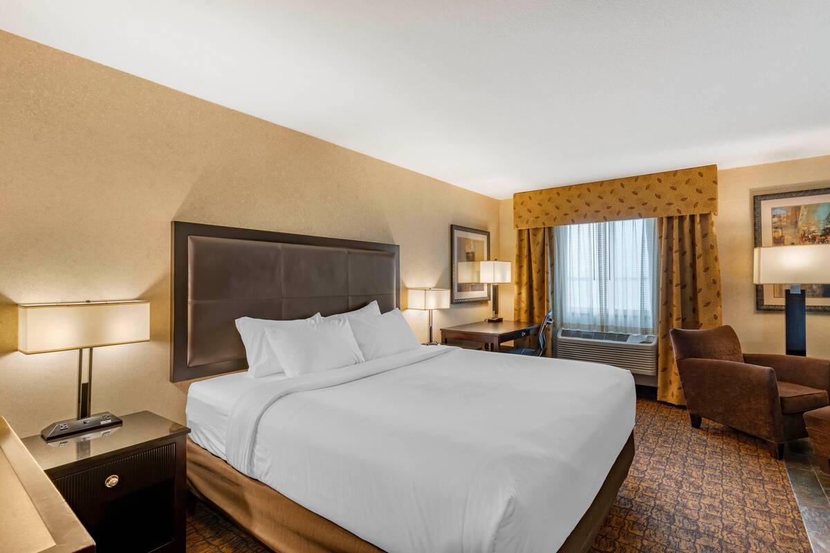 A hotel room with a large bed, two bedside tables with lamps, a chair, a desk, and a window with curtains.