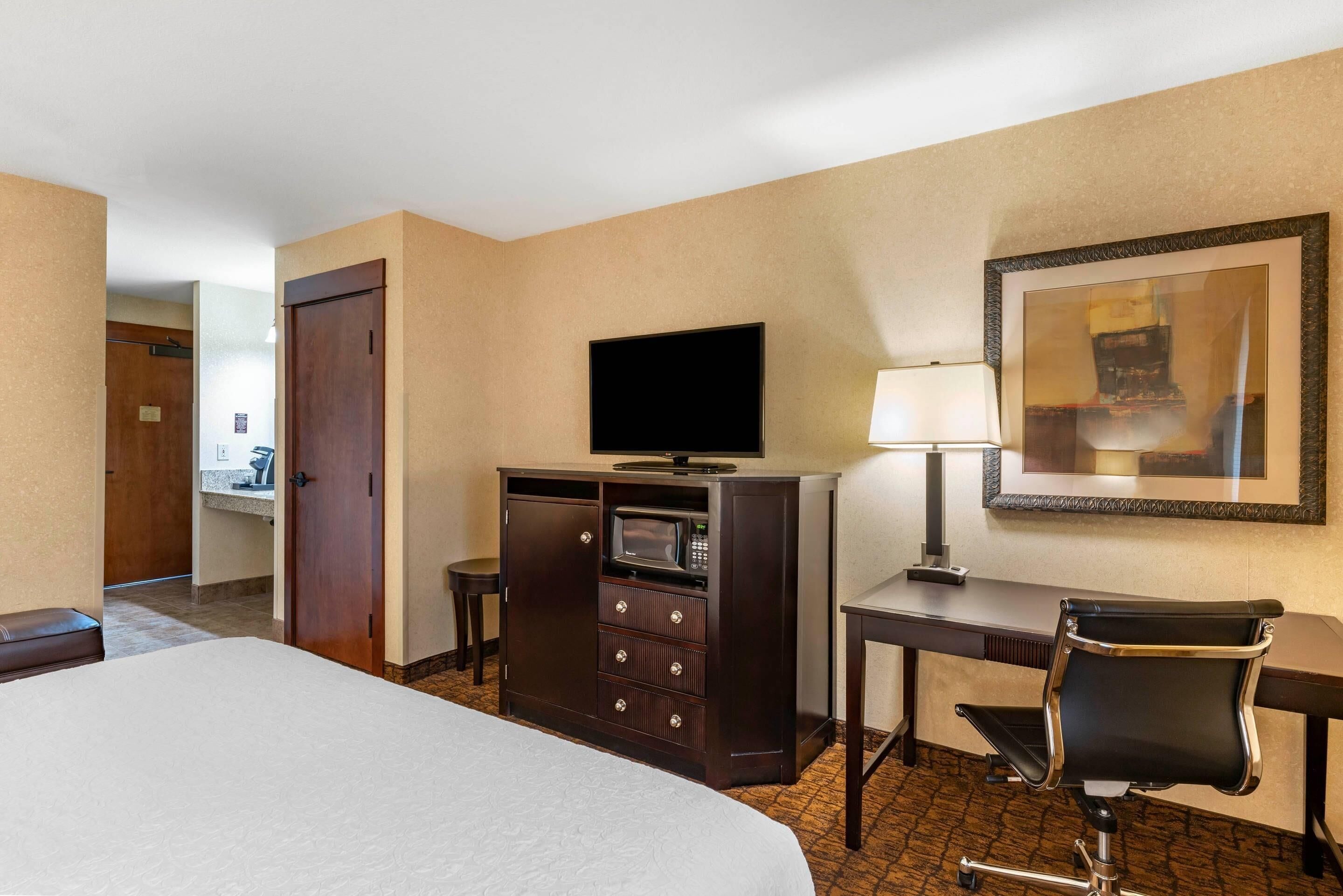 A hotel room with a bed, a desk with a lamp, a television, a minibar, and a chair.