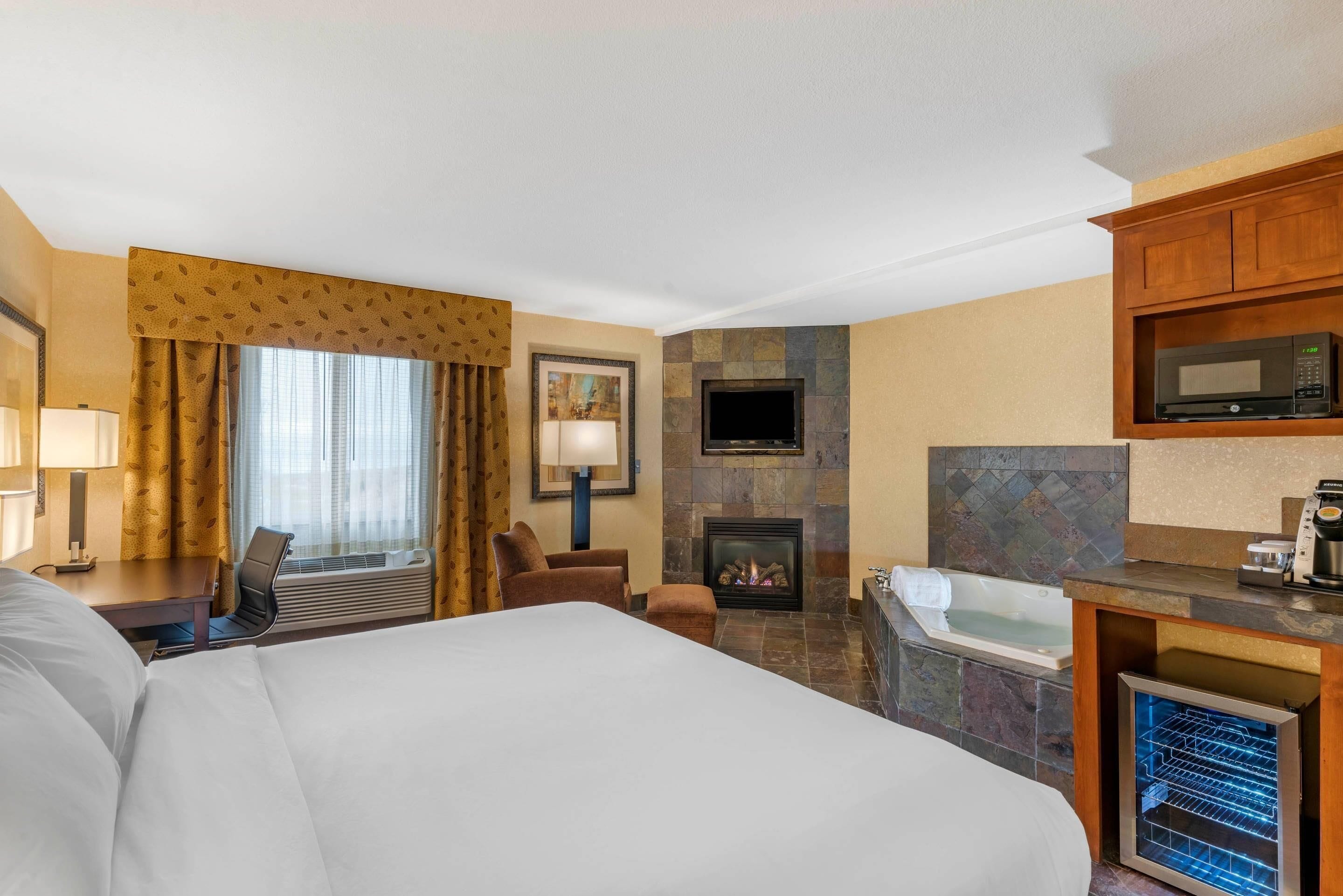 A hotel room with a large bed, a desk with a lamp, a chair, a fireplace, a television, a microwave, and a bathtub.