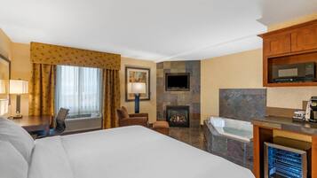 A hotel room with a large bed, a desk with a lamp, a chair, a fireplace, a television, a microwave, and a bathtub.
