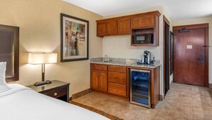 Habitación de hotel con cama, mesita de noche, lámpara, un cuadro, armarios de madera, microondas, refrigerador y lavavajillas integrado.