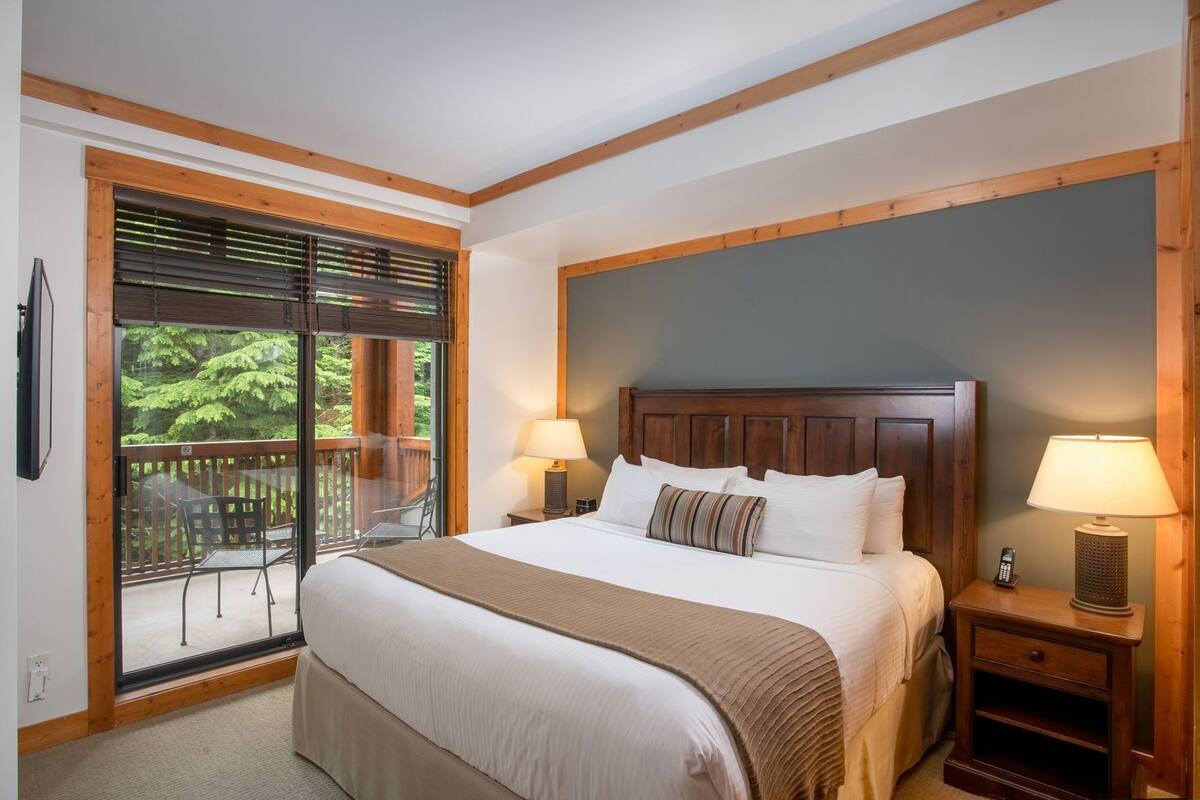 Hotelkamer met een groot bed, twee bedlampjes, een houten hoofdeinde, een balkon met uitzicht op bomen en een tafeltje met een lamp.