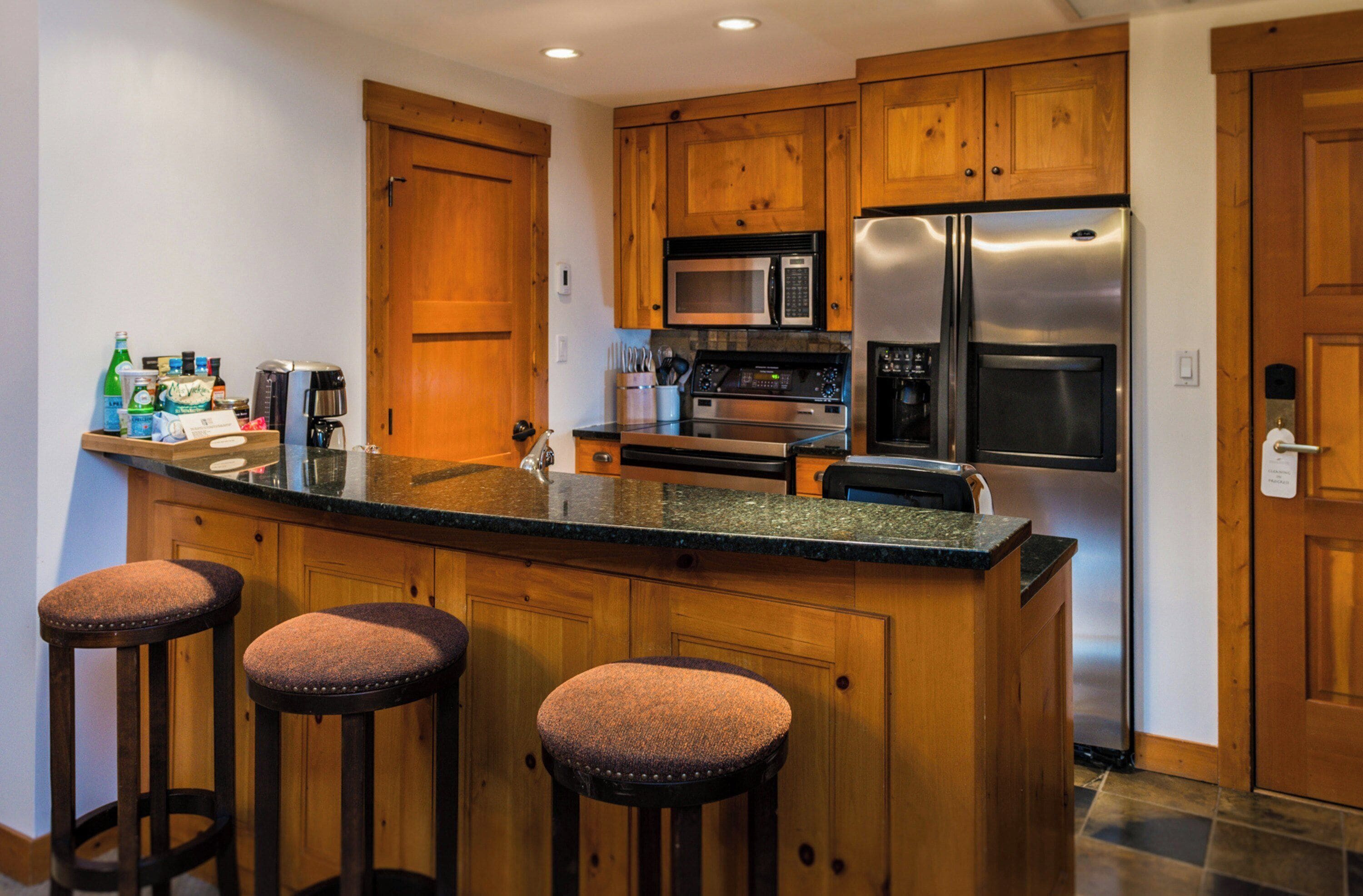 A kitchen with wooden cabinets, a stainless steel refrigerator, a microwave, and a coffee maker. There are three stools at the counter.