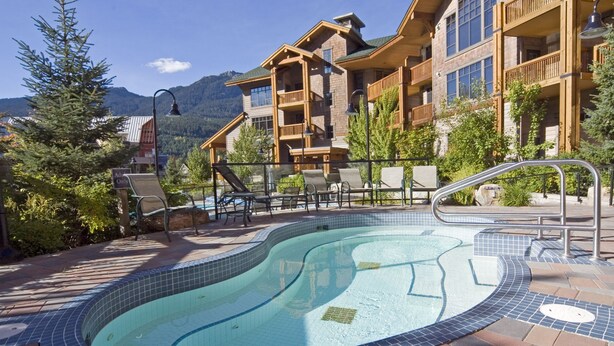 A pool area with a tiled edge, lounge chairs, and a building with balconies in the background. - First Tracks Lodge, by Lodging Ovations (Whistler)