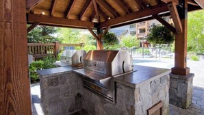 An outdoor stone barbecue area with a metal grill and a built-in oven, under a wooden pergola. - First Tracks Lodge, by Lodging Ovations (Whistler)