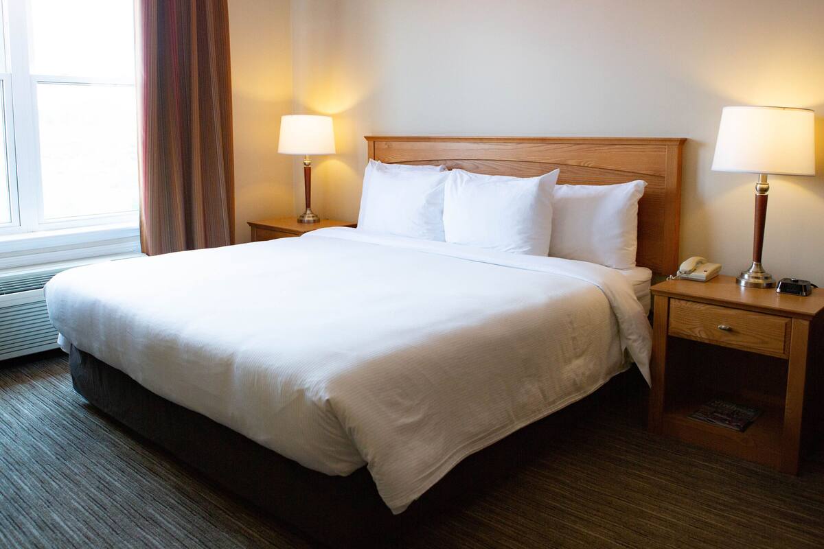 Una cama bien hecha con sábanas blancas, dos lámparas de noche, una mesita de madera y una ventana con cortinas.