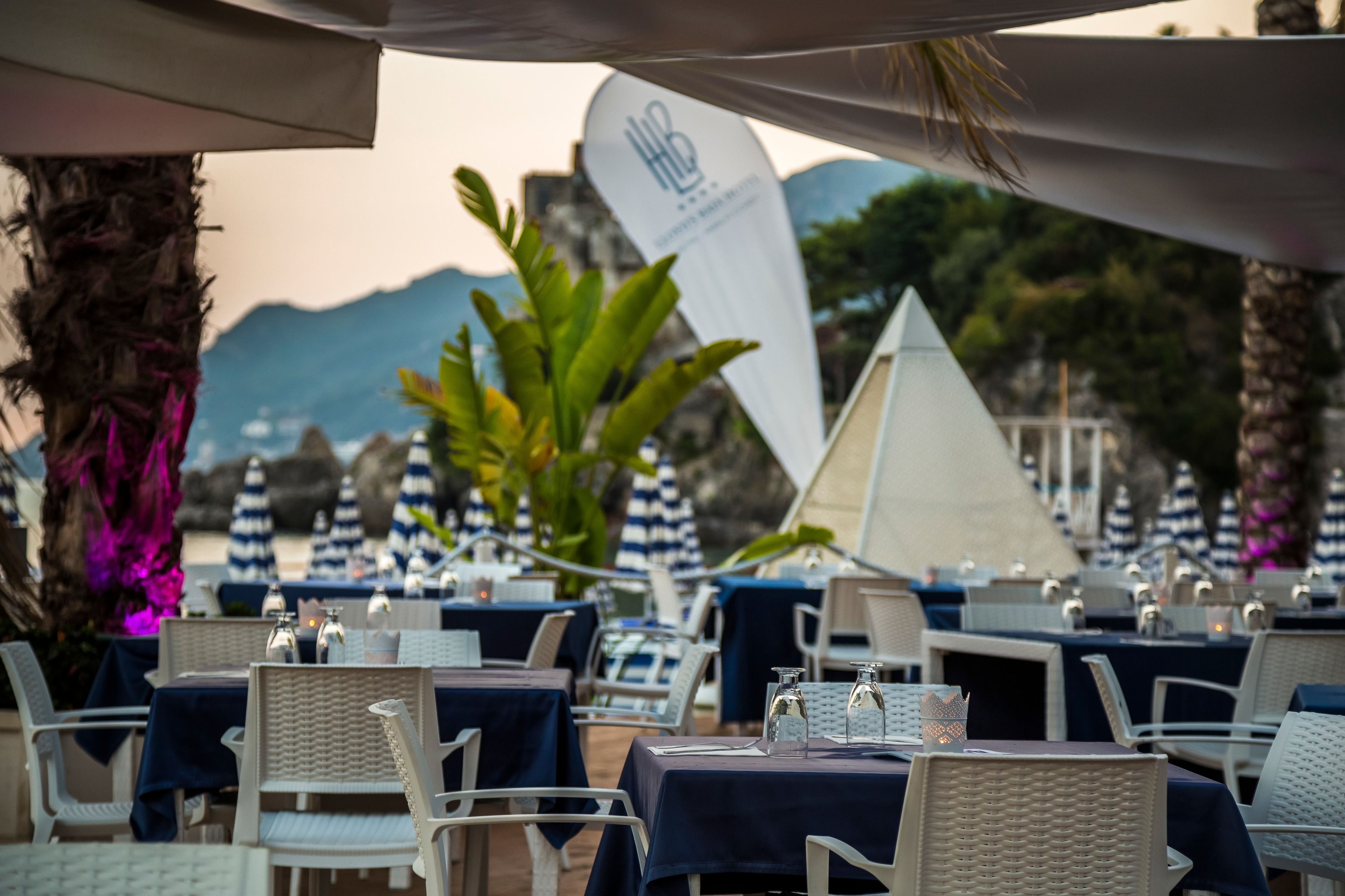 An outdoor dining area with tables and chairs, a large white banner, and a mountain view in the background.