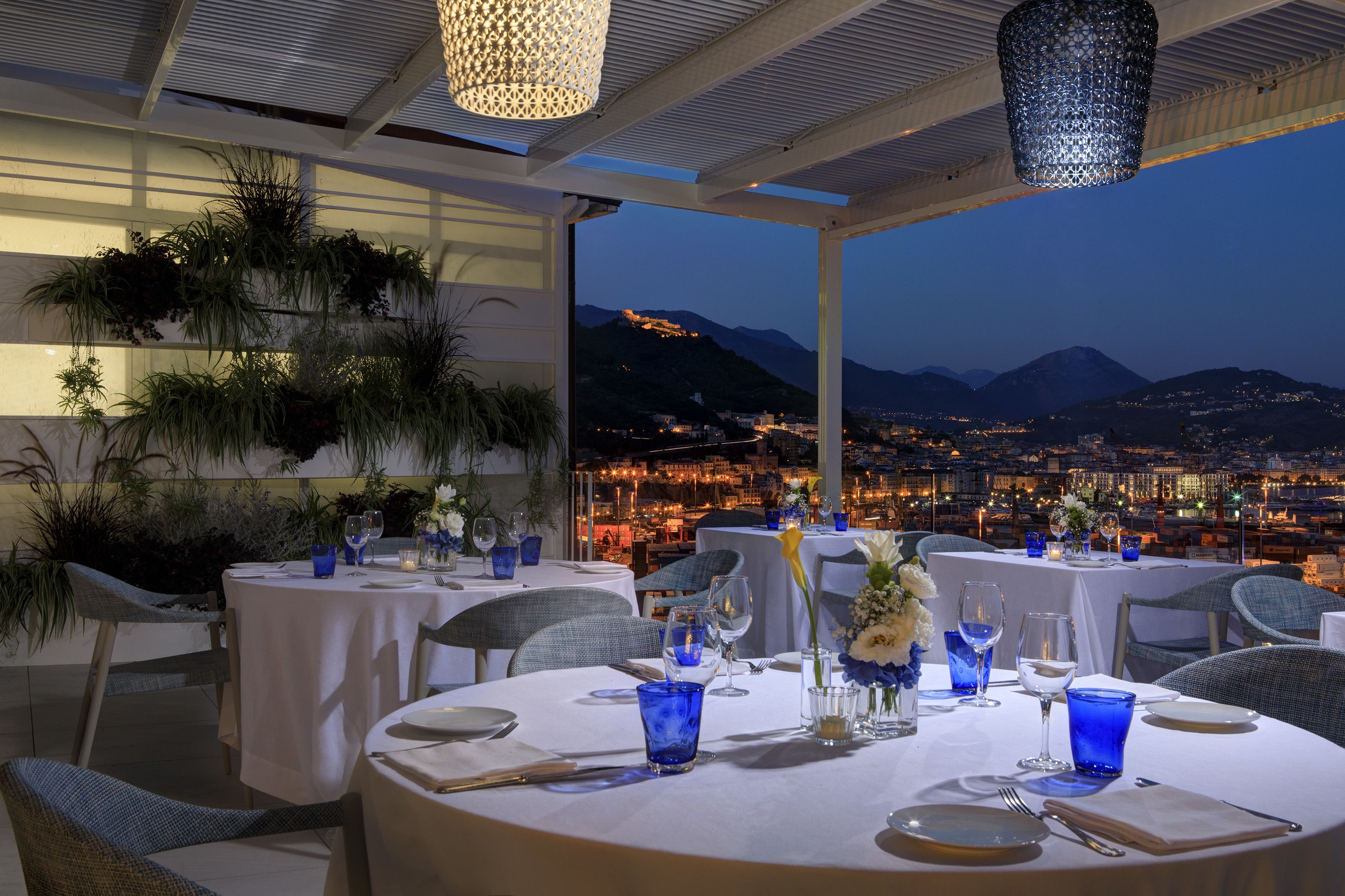 Un restaurante con mesas redondas preparadas para comer, con vasos de vidrio azul y manteles blancos.