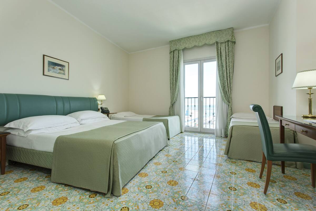 Habitació d'hotel amb llit, dues taules de nit, escriptori, cadira i finestra amb cortines.