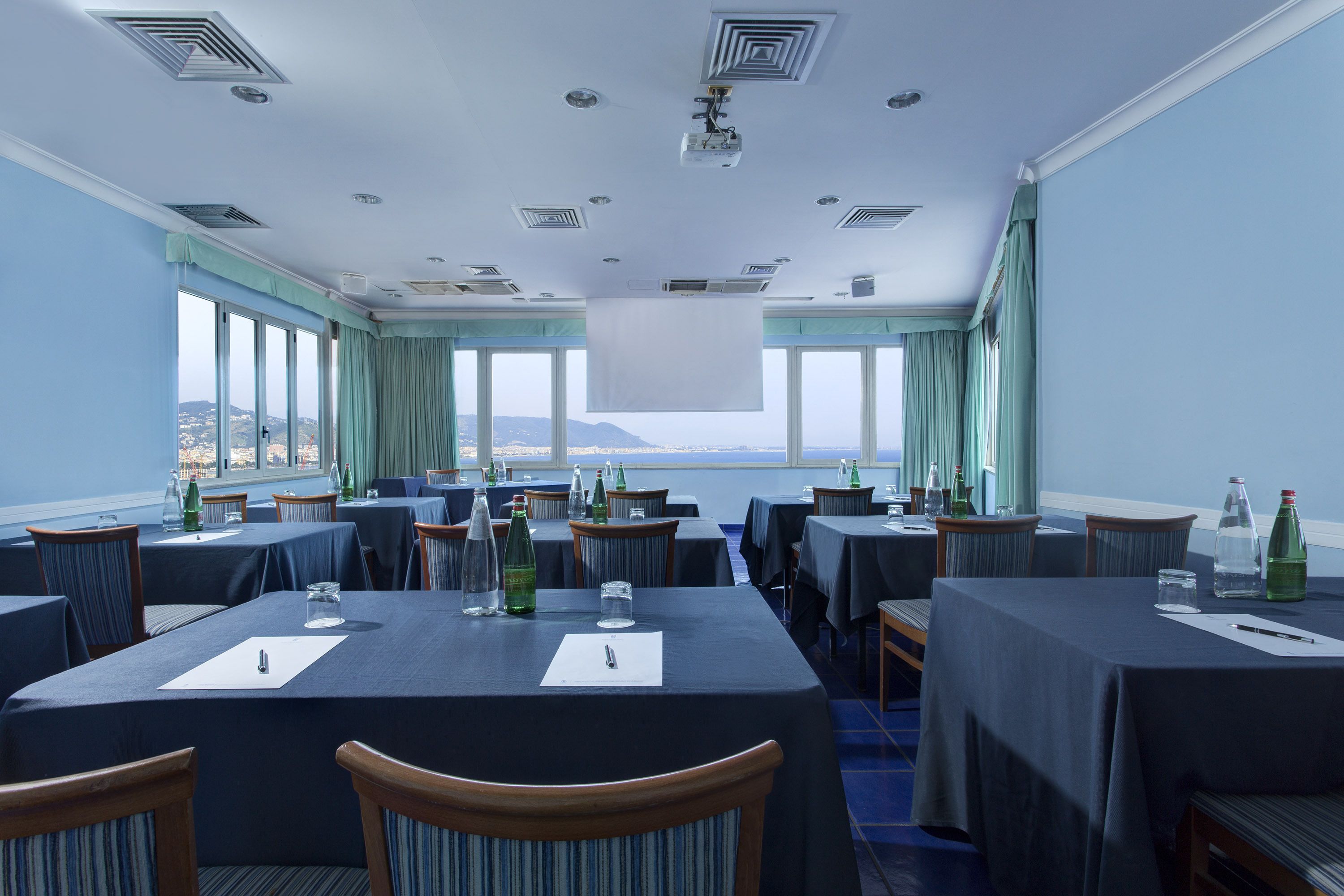 A conference room with tables set for meetings, large windows with curtains, and a view of the sea.