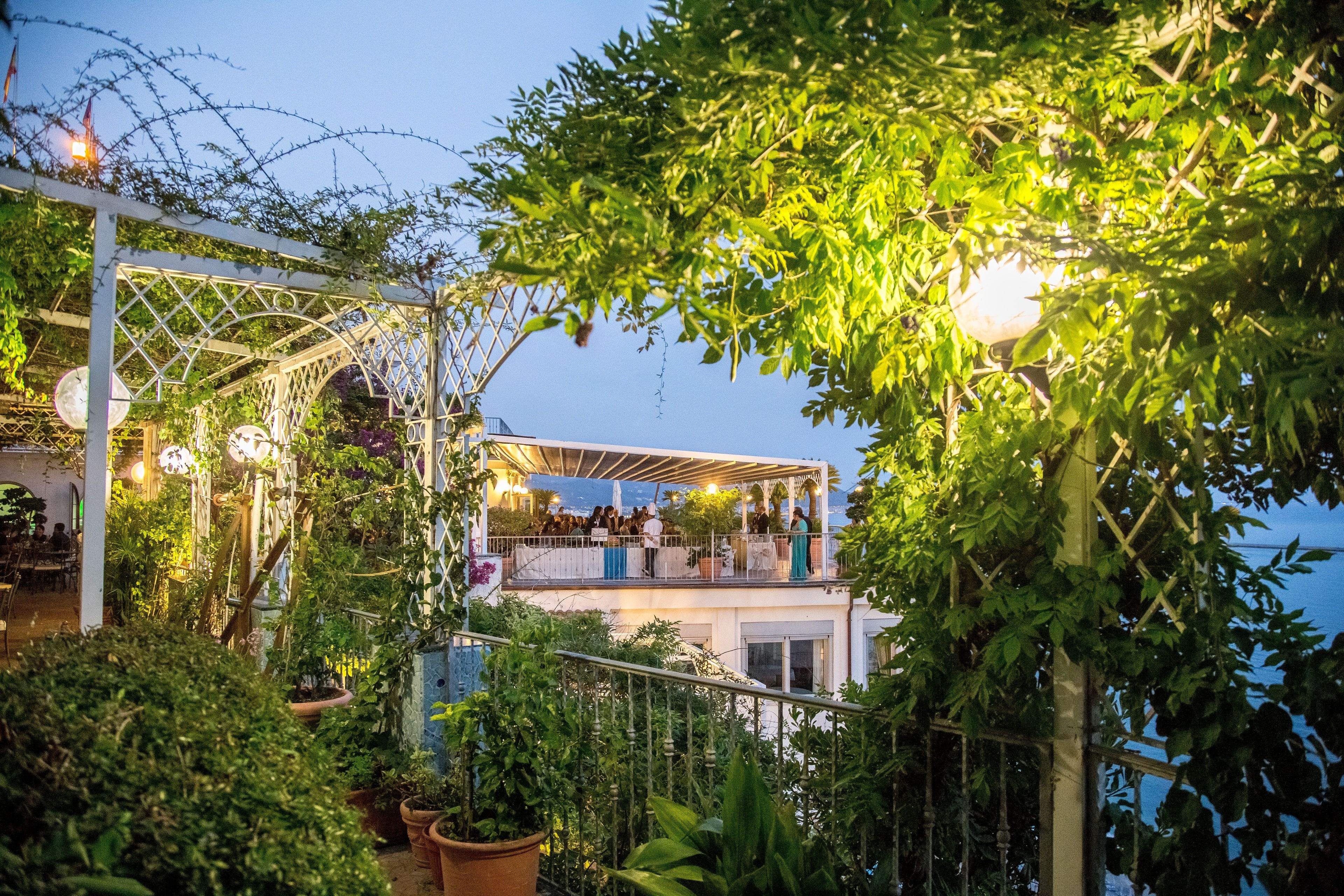 Una terraza exterior con una pérgola blanca, plantas en macetas y un área de piscina.