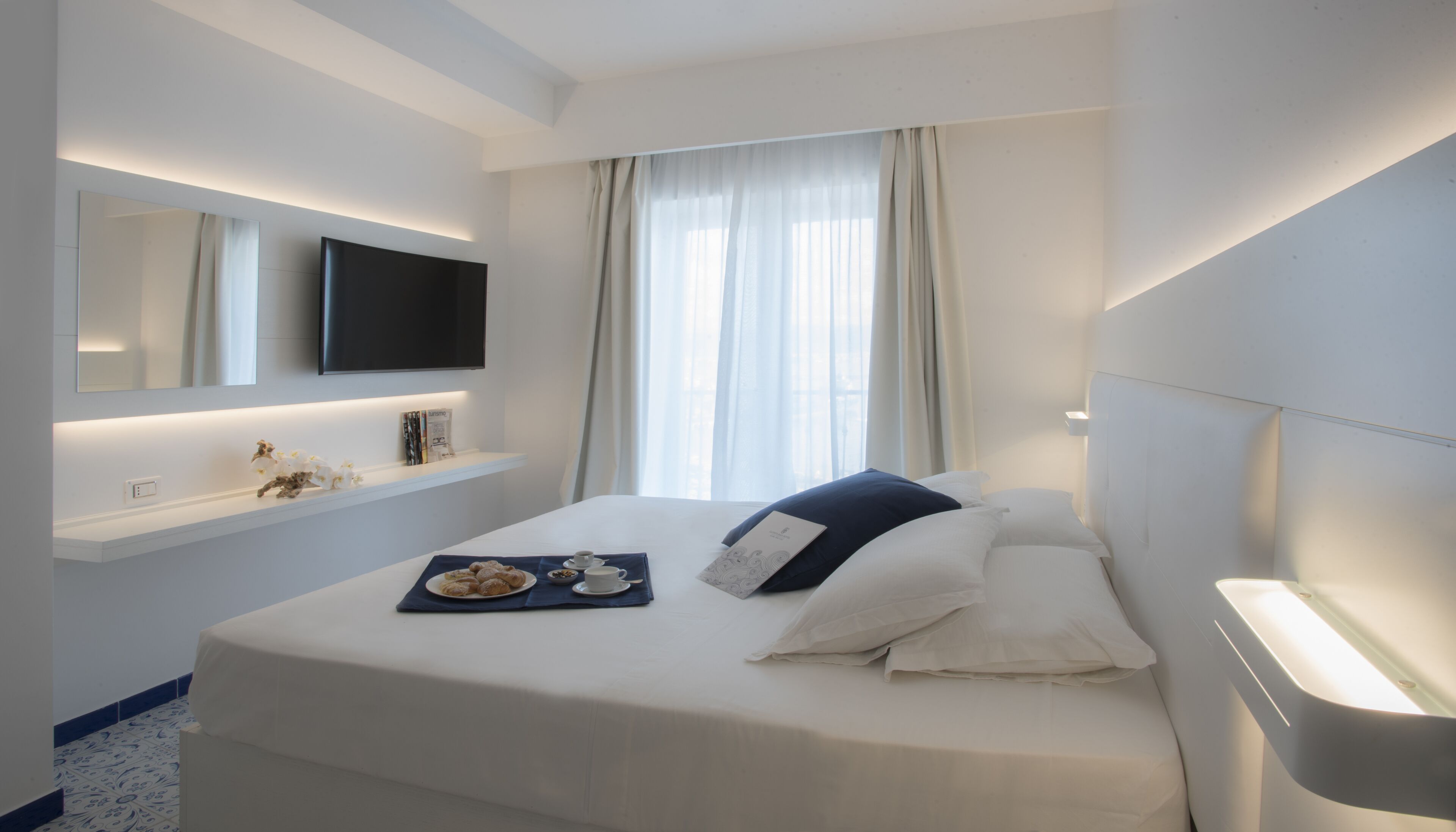 A hotel room with a bed, a flat-screen TV mounted on the wall, a bedside table with a tray of pastries, and a window with curtains.