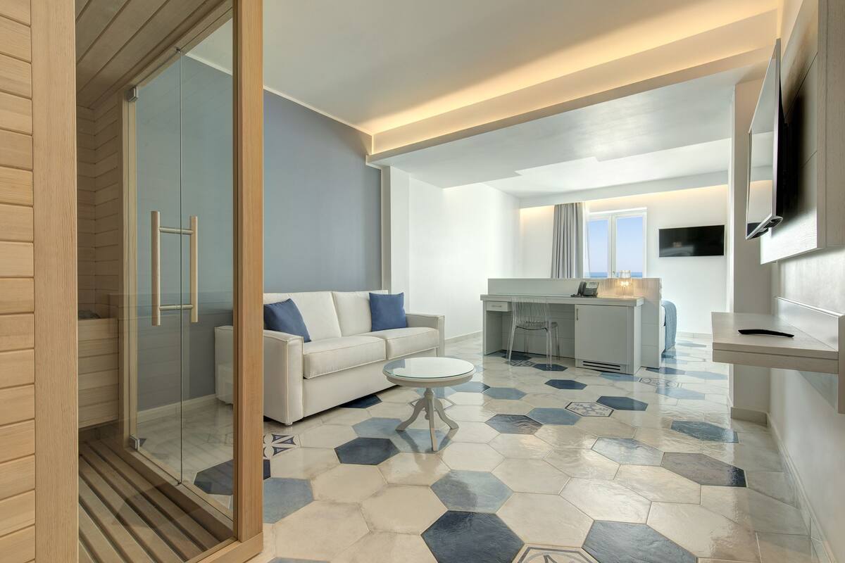 Un saló modern amb una dutxa tancada amb vidre, un sofà blanc amb coixins blaus, una taula de centre rodona i un paviment de disseny hexagonal.